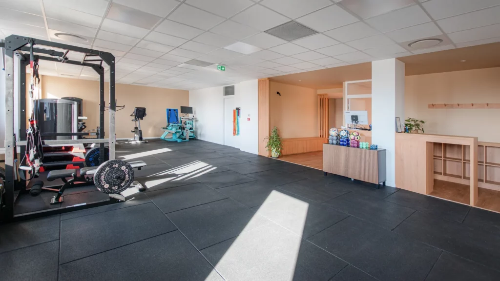Large vue d'intérieur du centre Hélios Santé à Nantes montrant le plateau technique de sport-santé, le rack de musculation, les machines de cardio et l'espace d'accueil en bois. Sol sportif noir et éclairage naturel.