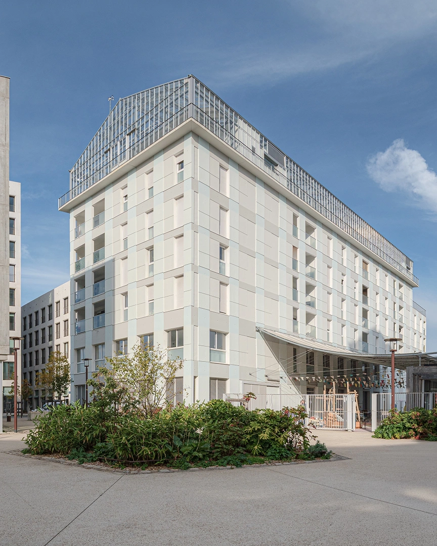 Une photo d'angle en plein jour du bâtiment principal des 5 Ponts à Nantes. La structure comporte une base en panneaux de béton clairs avec loggias, surmontée d'une grande serre vitrée sur deux niveaux. Un aménagement paysager avec arbustes est visible au premier plan sur une place en béton désactivé.