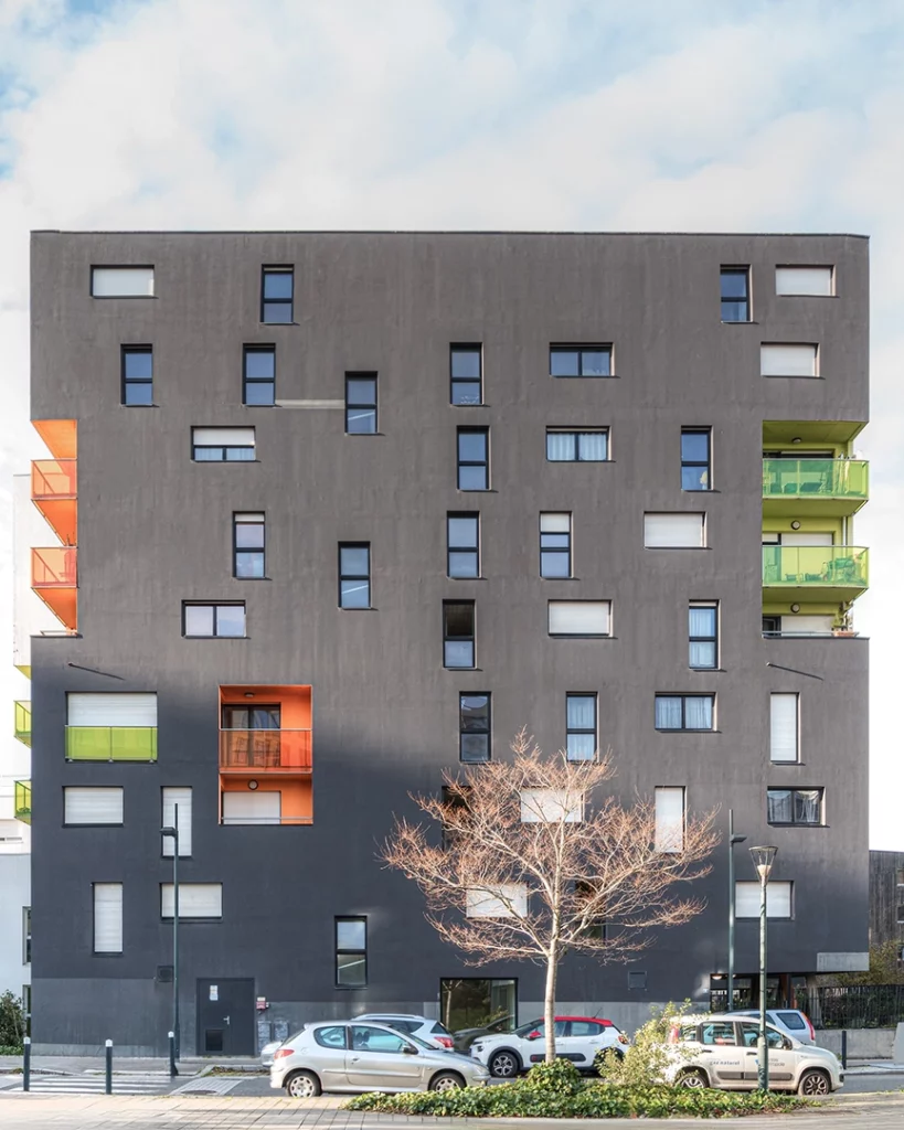 Façade grise foncée du projet Chroma à Nantes avec une disposition irrégulière des fenêtres et des balcons encastrés orange et verts.