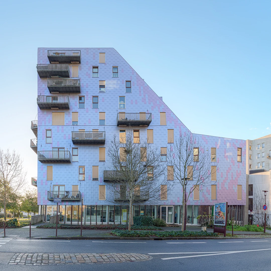 Façade latérale de l'immeuble Le Ruban à Nantes montrant sa forme asymétrique, ses balcons empilés et ses larges baies vitrées au rez-de-chaussée, sous un ciel clair.