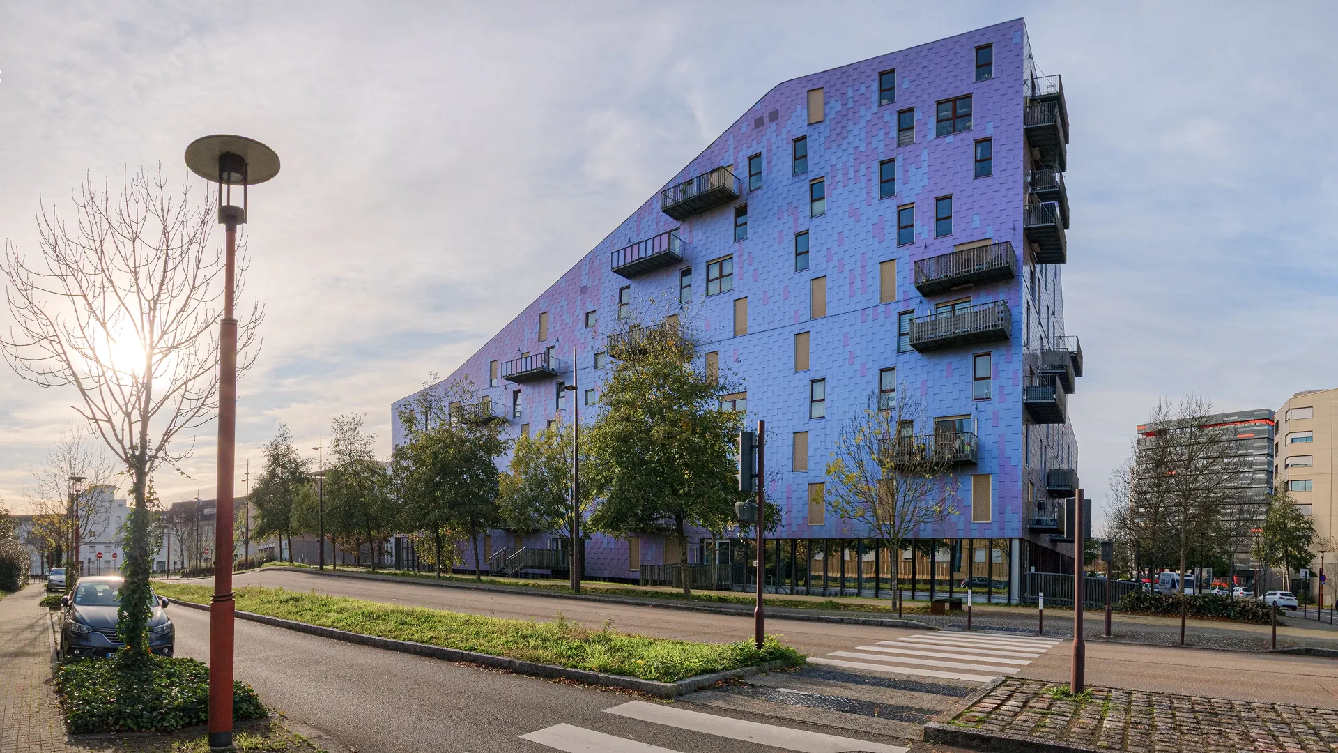 Vue en perspective de l'immeuble Le Ruban à Nantes depuis la rue, montrant sa façade texturée violette, sa toiture en pente et ses balcons métalliques en saillie.
