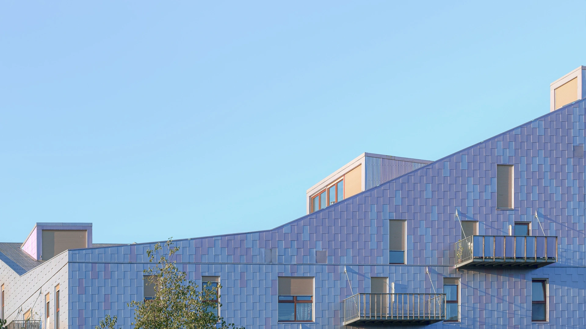 Détail de la ligne de toiture en pente de l'immeuble Le Ruban à Nantes, montrant les émergences des derniers étages et le bardage métallique bleu et violet sur fond de ciel clair.