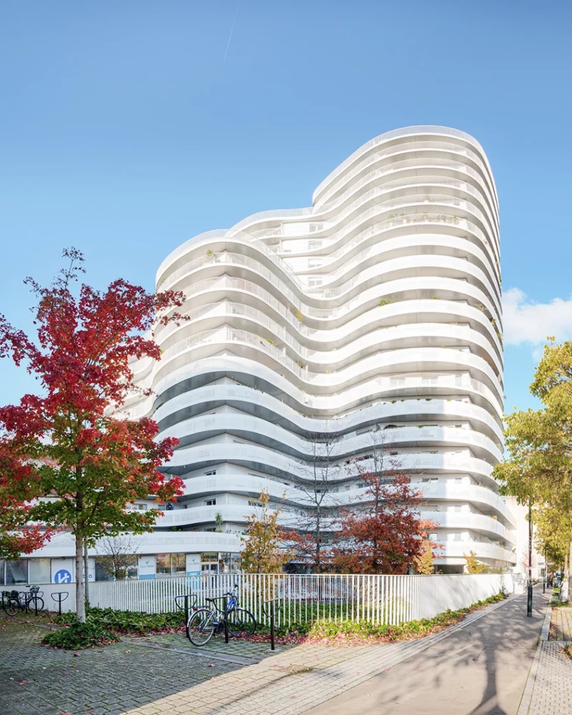 Façade blanche aux lignes courbes de l'immeuble New'R à Nantes, capturée en contre-plongée avec un érable rouge vif au premier plan sous un ciel bleu clair.