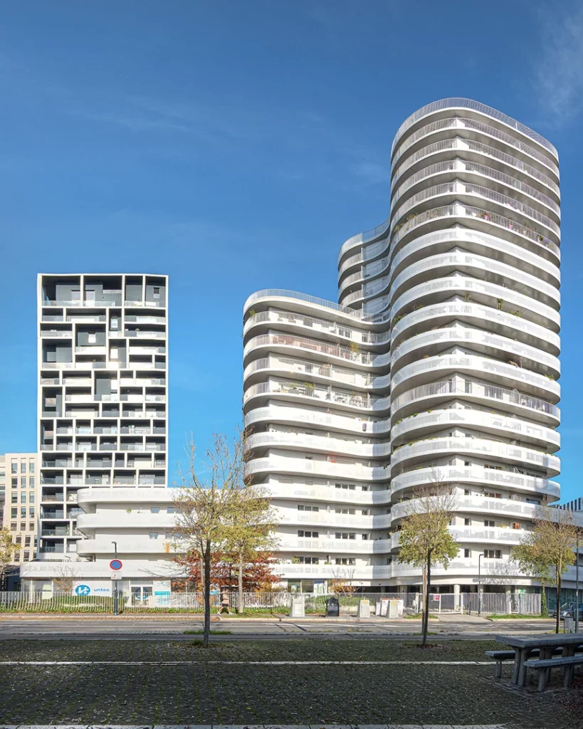 Comparaison visuelle entre les courbes blanches organiques de l'immeuble New'R et la structure cubique sombre de son voisin Hémistone à Nantes, sous un ciel bleu intense.