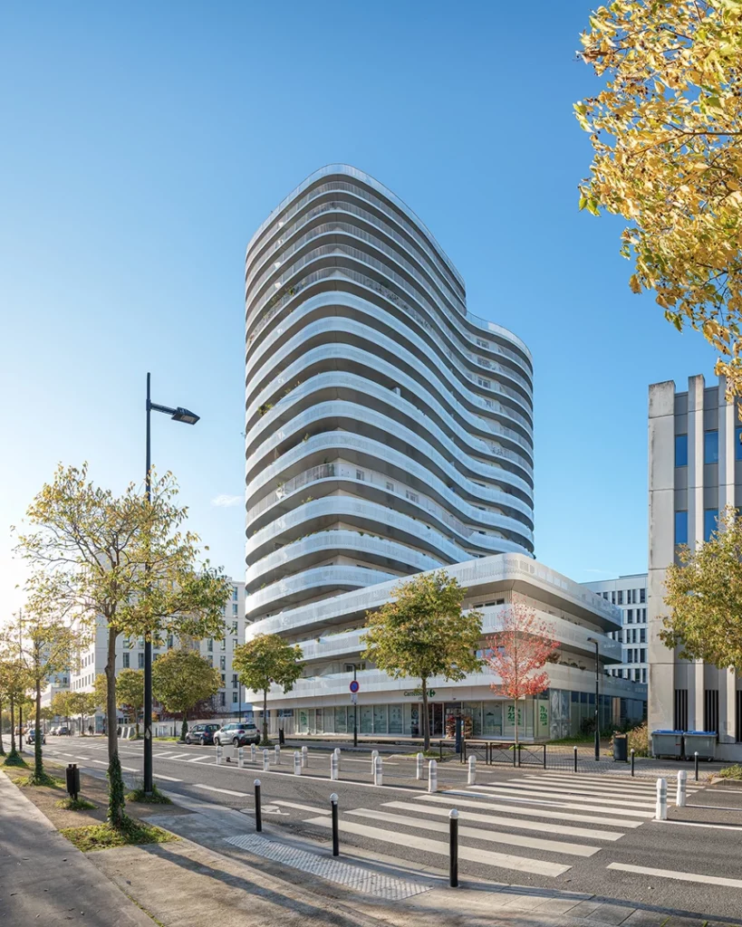 Vue d'ensemble de la tour New'R à Nantes depuis le boulevard, montrant sa silhouette élancée derrière des arbres d'alignement, un passage piéton et les bâtiments tertiaires environnants.