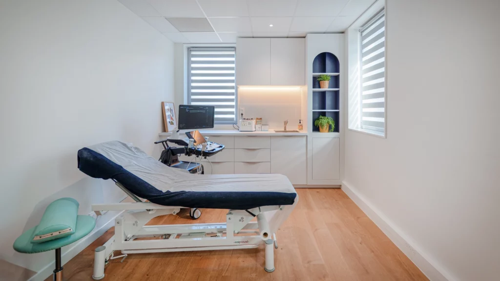 Photographie d'intérieur d'une salle de consultation médicale au cabinet Hélios Santé à Nantes, avec table d'examen ergonomique, appareil d'échographie, mobilier blanc design et parquet clair.