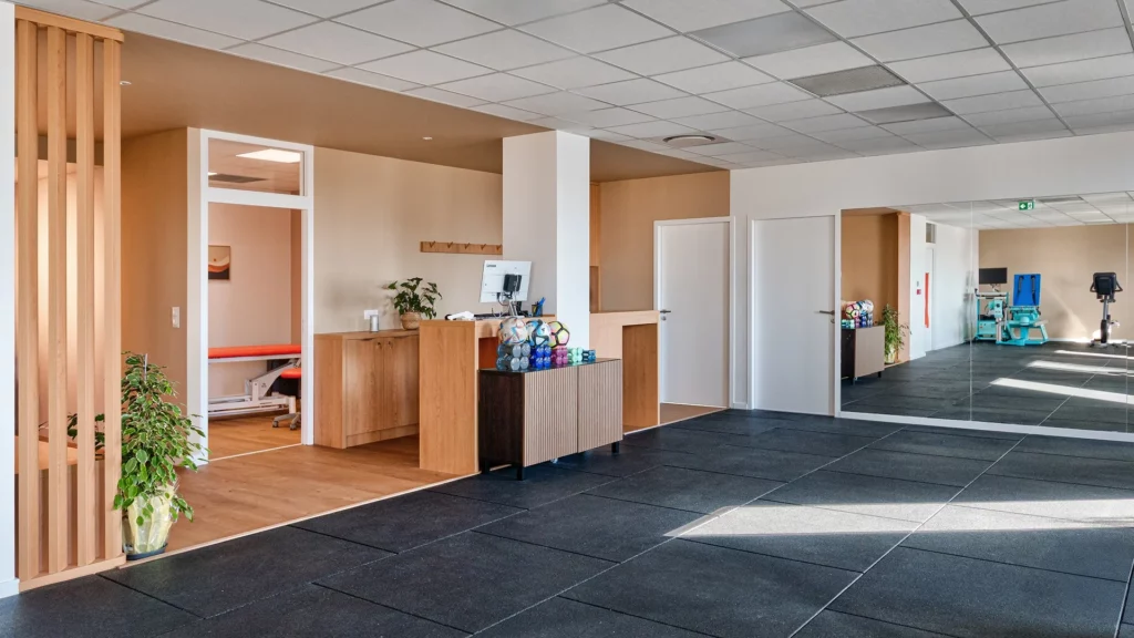 Vue d'ensemble de l'intérieur du cabinet Hélios Santé à Nantes. On y voit un comptoir en bois clair, une cabine de soin avec table de massage, un grand miroir et le plateau de rééducation. Design moderne avec claustra en bois et plantes vertes.