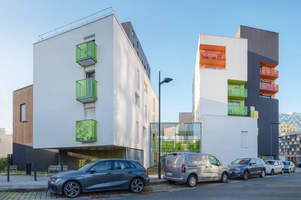Vue sur deux bâtiments du projet Chroma à Nantes reliés par une structure vitrée, avec des balcons saillants verts et orange et des voitures stationnées au premier plan.