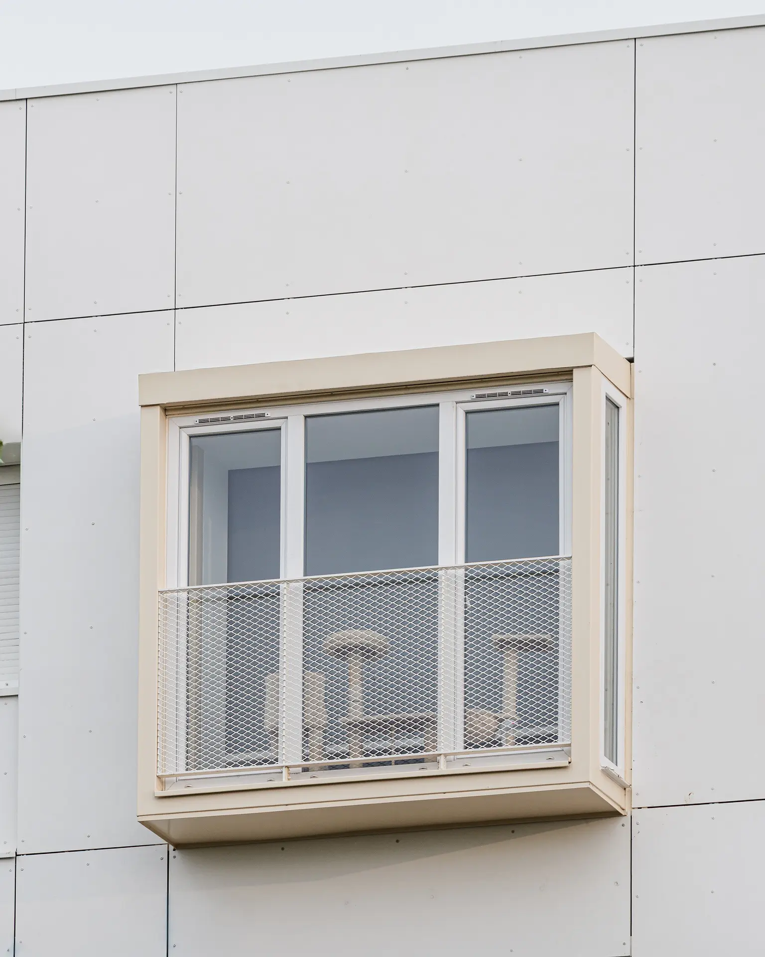 Gros plan sur un bow-window à structure beige et garde-corps en maille métallique fine, installé sur une façade blanche isolée.
