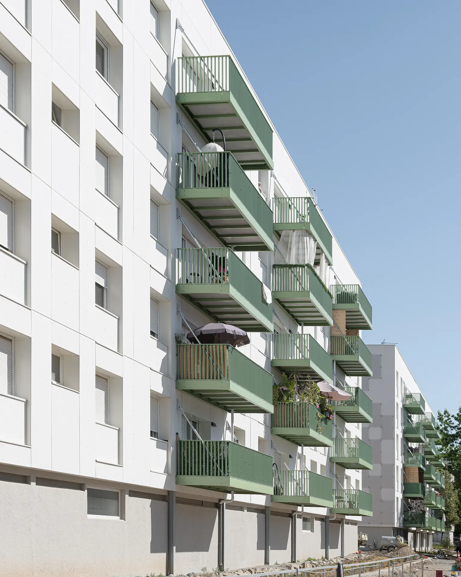 Vue en perspective d'une longue façade de logements sociaux habillée de multiples balcons métalliques d'un vert sapin contemporain.